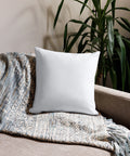 White 18 by 18 inch premium throw pillow showing its plain white back, on a sofa with a knitted throw beside a leafy plant.