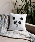 White 18 by 18 inch premium throw pillow with a black ink-splatter panda face on the front, on a sofa with a knitted throw beside a leafy plant.