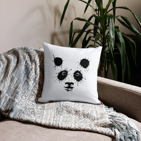 White 18 by 18 inch premium throw pillow with a black ink-splatter panda face on the front, on a sofa with a knitted throw beside a leafy plant.
