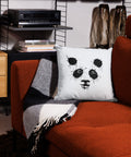 White 18 by 18 inch premium throw pillow with a black ink-splatter panda face on the front, on a rust-coloured sofa.