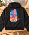 Black heavyweight hoodie with colourful Antidepresantai cat jar front print, laid on an oak bench beside a linen cushion and houseplant.