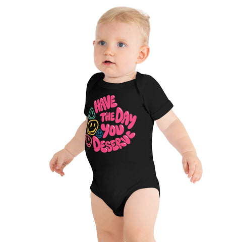 Baby model wearing a black short-sleeve Have the Day You Deserve bodysuit with one large pink word design and colourful smiley faces printed high on the front.