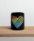 Rainbow Heart 11 oz black glossy ceramic mug on a light wooden serving tray against a pale grey wall, showing its single colourful heart front print.