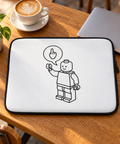 White brick figure laptop sleeve on a sunlit oak café table beside a silver laptop, coffee and green plant.