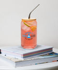 Alcohol Is a Solution 473 ml can-shaped glass with bamboo lid and metal straw, filled with a pink citrus drink on stacked books, showing its single blue-and-black front print.
