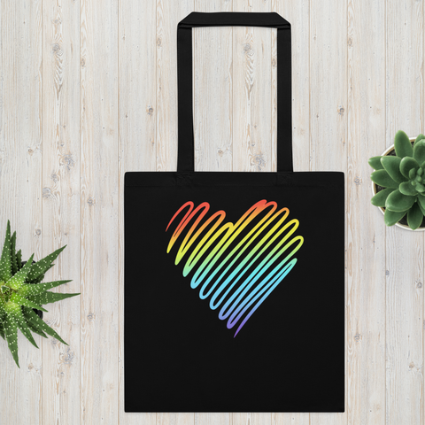 Black Rainbow Heart cotton tote bag on pale wooden boards with succulents, showing one large colourful heart on the front and long black handles.