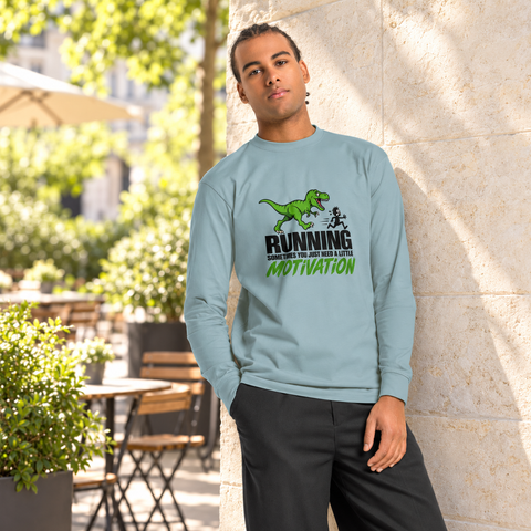 Model wearing the Agave Dinosaur Motivation men’s heavyweight long-sleeve tee on a sunlit cafe terrace beside a pale stone wall and greenery, showing its single front print.