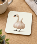 Glossy cream cork-back coaster with a surreal garlic swan illustration, on an oak table beside coffee and linen.