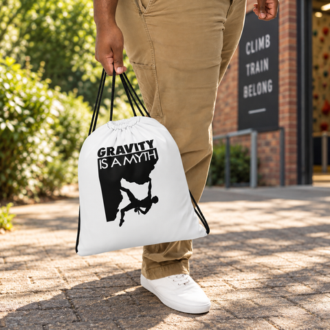 White Gravity Is a Myth drawstring bag carried along a sunlit path outside a climbing gym, with its single black front design visible.