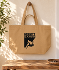 Latte Gravity Is a Myth recycled cotton shopping bag hanging in a sunlit entryway above a wooden bench beside a green plant, showing its single black front print.