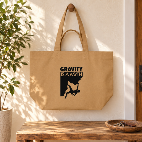Latte Gravity Is a Myth recycled cotton shopping bag hanging in a sunlit entryway above a wooden bench beside a green plant, showing its single black front print.