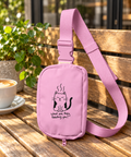 Pink Grumpy Cat sling bag with its adjustable strap on a sunlit wooden cafe bench beside coffee and a green plant.