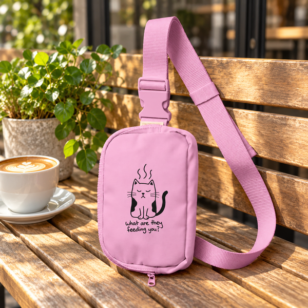 Pink Grumpy Cat sling bag with its adjustable strap on a sunlit wooden cafe bench beside coffee and a green plant.