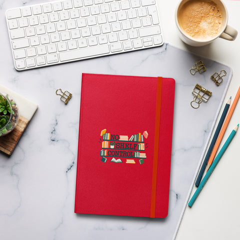 Red No Shelf Control hardcover lined notebook on a pale marble desk beside a white keyboard, coffee, pencils and binder clips, showing one complete bookish front-cover design.