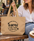 Woman in a white blouse holding a Latte recycled cotton shopping bag with the black Karma quote design on a wooden outdoor cafe bench.