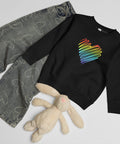 Black Rainbow Heart kids’ crew neck sweatshirt laid beside patterned jeans and a soft toy rabbit, showing its single colourful front print.