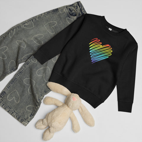 Black Rainbow Heart kids’ crew neck sweatshirt laid beside patterned jeans and a soft toy rabbit, showing its single colourful front print.