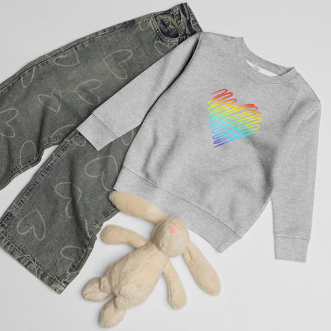 Heather Rainbow Heart kids’ crew neck sweatshirt laid beside patterned jeans and a soft toy rabbit, showing its single colourful front print.