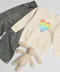 Natural Heather Rainbow Heart kids’ crew neck sweatshirt laid beside patterned jeans and a soft toy rabbit, showing its single colourful front print.