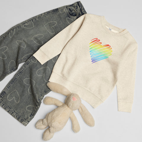 Natural Heather Rainbow Heart kids’ crew neck sweatshirt laid beside patterned jeans and a soft toy rabbit, showing its single colourful front print.