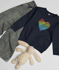 Navy Rainbow Heart kids’ crew neck sweatshirt laid beside patterned jeans and a soft toy rabbit, showing its single colourful front print.