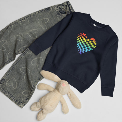 Navy Rainbow Heart kids’ crew neck sweatshirt laid beside patterned jeans and a soft toy rabbit, showing its single colourful front print.