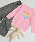 Pink Rainbow Heart kids’ crew neck sweatshirt laid beside patterned jeans and a soft toy rabbit, showing its single colourful front print.