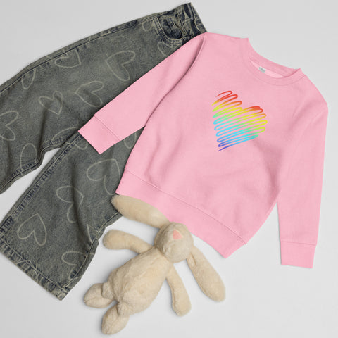 Pink Rainbow Heart kids’ crew neck sweatshirt laid beside patterned jeans and a soft toy rabbit, showing its single colourful front print.
