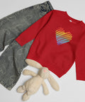 Red Rainbow Heart kids’ crew neck sweatshirt laid beside patterned jeans and a soft toy rabbit, showing its single colourful front print.
