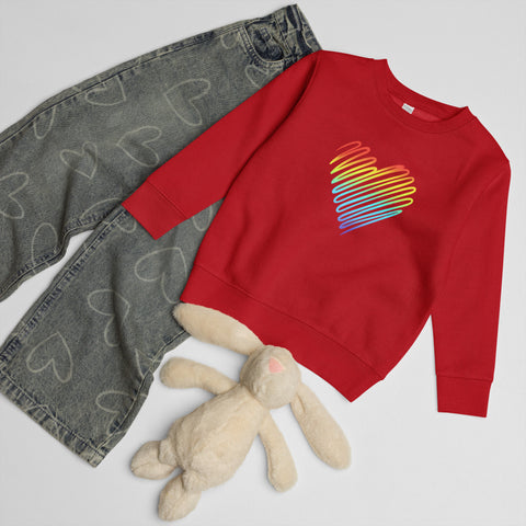 Red Rainbow Heart kids’ crew neck sweatshirt laid beside patterned jeans and a soft toy rabbit, showing its single colourful front print.