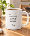 White 11 oz ceramic mug with a Lithuanian cat humour illustration, on a sunny oak table beside coffee and a sleeping cat.