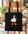 Black cotton tote with a cheeky white goose graphic hanging on a wooden café chair beside coffee and a book.