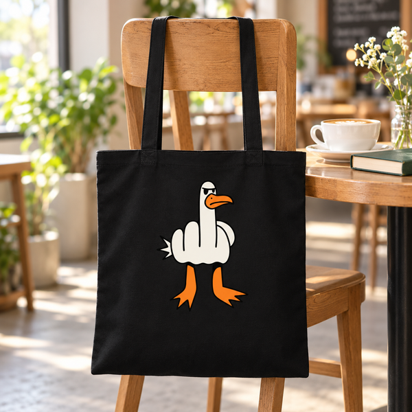Black cotton tote with a cheeky white goose graphic hanging on a wooden café chair beside coffee and a book.