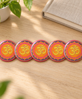 Five matching Vibrant Om Mandala 1.25-inch glossy pin buttons on a pale oak desk beside a linen notebook and green plant, each showing one complete colourful Om front design.