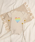 The organic natural Rainbow Heart organic cotton baby bodysuit laid on cream bedding.