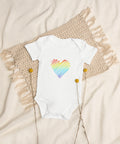 The white Rainbow Heart organic cotton baby bodysuit laid on cream bedding.