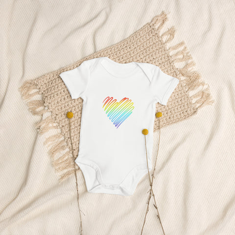 The white Rainbow Heart organic cotton baby bodysuit laid on cream bedding.