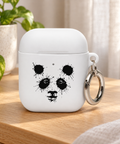 White Ink Splatter Panda rubber case for AirPods 1st and 2nd generation on a light oak bedside table beside a green plant and linen curtain, showing its single black panda front print and metal carabiner.