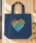 Denim Blue Rainbow Heart organic cotton tote bag hanging from a wooden peg in a sunlit home entryway beside a green plant, showing one complete rainbow heart front print.
