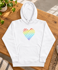 White Rainbow Heart pullover hoodie laid on an oak bench in a sunlit studio beside a green plant and canvas sneakers.
