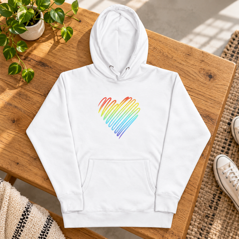 White Rainbow Heart pullover hoodie laid on an oak bench in a sunlit studio beside a green plant and canvas sneakers.