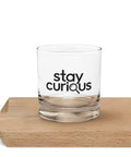 Clear Stay Curious rocks glass with a single black magnifying-glass lettering design on the front, standing on a wooden board.