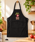 Black Rooting for Dinner organic cotton apron hanging in a sunlit kitchen beside vegetables, plants and wooden utensils.