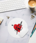 Pierced Skull Heart round mouse pad on a pale marble desk beside a keyboard, coffee and coloured pencils, showing one tattoo-style design and a curled edge.