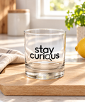 Clear Stay Curious rocks glass on a wooden board in a sunlit kitchen beside a linen cloth, lemon and green plant.