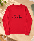 Red Stay Curious youth long-sleeve tee laid on a sunlit wooden study desk beside coloured pencils, a notebook and a green plant.