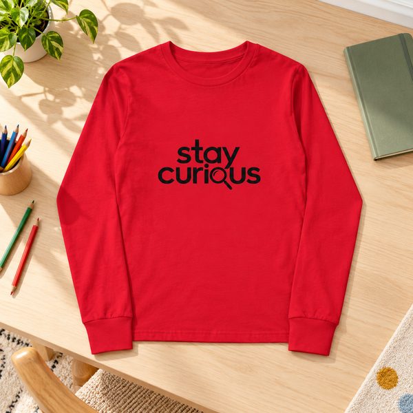 Red Stay Curious youth long-sleeve tee laid on a sunlit wooden study desk beside coloured pencils, a notebook and a green plant.