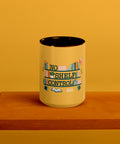 15 oz white No Shelf Control ceramic mug with golden yellow interior, rim and handle, front view on a wooden board; book illustration and green lettering printed once on the front.