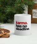 White 11 oz Karma Has No Deadline ceramic mug beside pine branches, dried citrus and cinnamon, showing the full red-and-black quote on the front.