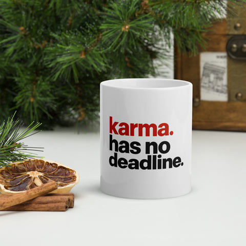 White 11 oz Karma Has No Deadline ceramic mug beside pine branches, dried citrus and cinnamon, showing the full red-and-black quote on the front.